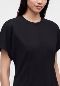 Femme portant un haut noir uni à manches courtes avec un col rond et des détails discrets de coutures à l'avant sur fond clair.