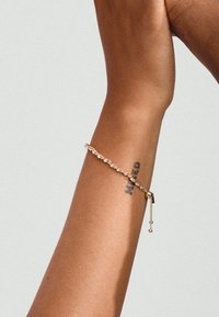 Bracelet doré orné de cristaux transparents, avec un design de chaîne délicat et un petit accent de perle suspendue, porté sur un poignet portant un tatouage.
