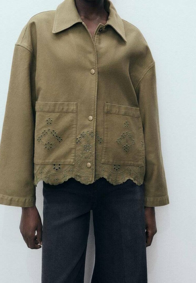 Personne portant une veste boutonnée vert olive avec deux grandes poches avant et une broderie florale en œillets au-dessus de l'ourlet festonné.