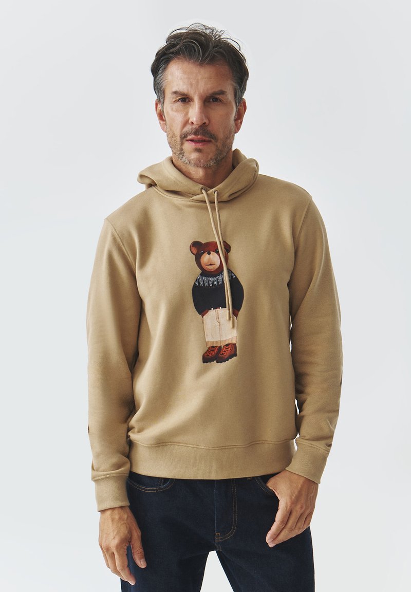Sudadera color caqui con cordón, con un gráfico de un oso en un suéter azul, combinada con pantalones beige claros y zapatos marrones. Textura suave.