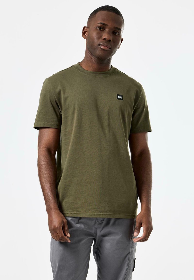 Tricou din bumbac de culoare verde măsliniu, cu guler rotund și mâneci scurte; are o etichetă mică neagră pe piept. Asortat cu pantaloni cargo gri.