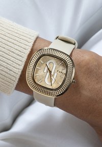 Reloj con correa de silicona beige, caja cuadrada en tono dorado, esfera con relieve y bisel texturizado, que muestra el nombre de la marca "Furla."