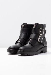 Botas de cuero negro hasta el tobillo con cremalleras plateadas, dos correas anchas y grandes hebillas plateadas sobre una superficie blanca.
