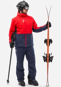 Veste de ski rouge et marine avec capuche, pantalon de ski marine, casque noir et lunettes de ski. Tient des skis avec des détails orange et des fixations noires.
