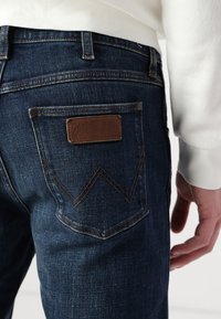 Jeans de mezclilla azul con un parche de cuero marrón que lleva el texto "Wrangler", equipados con costuras decorativas y remaches de metal.