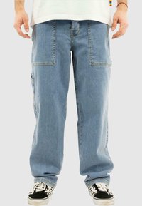 Lysblå bredbenspants lavet af denim, med store forlommer og en afslappet pasform. Båret med sorte og hvide sneakers.