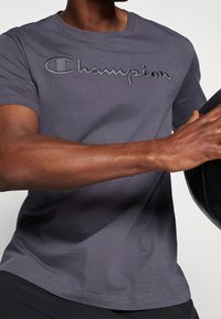 Camiseta de algodón gris con un logo bordado de "Champion" en un tono contrastante, de manga corta, cuello redondo y un ajuste relajado.