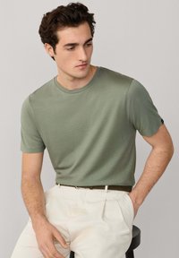 Junger Mann, der auf einem Hocker sitzt, trägt ein grünes Kurzarmhemd und weiße plissierte Hose, die Hände in den Taschen, schaut nach links unten.