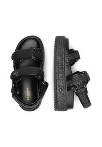 Sandalias negras con dos correas ajustables, textura tejida, un acabado brillante y una suela gruesa y texturada. Acentos dorados en las correas.
