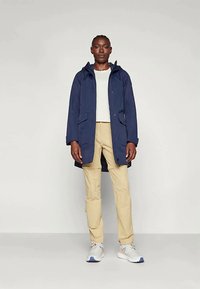 Tumši zilii ar kapuci mētelis ar rāvējslēdzēju, divām priekšējām kabatām un brīvu piegriezumu, apvienots ar gaiši beigiem biksēm un baltajiem sporta apaviem. Gluda auduma struktūra.