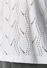 Tissu blanc avec un motif ondulé et des détails en œillets ouverts. La texture semble légère et respirante, avec une finition lisse.