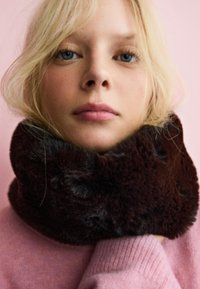 Col montant en fausse fourrure marron avec une texture douce, porté sur un pull rose clair. L'arrière-plan est d'une couleur rose uni.