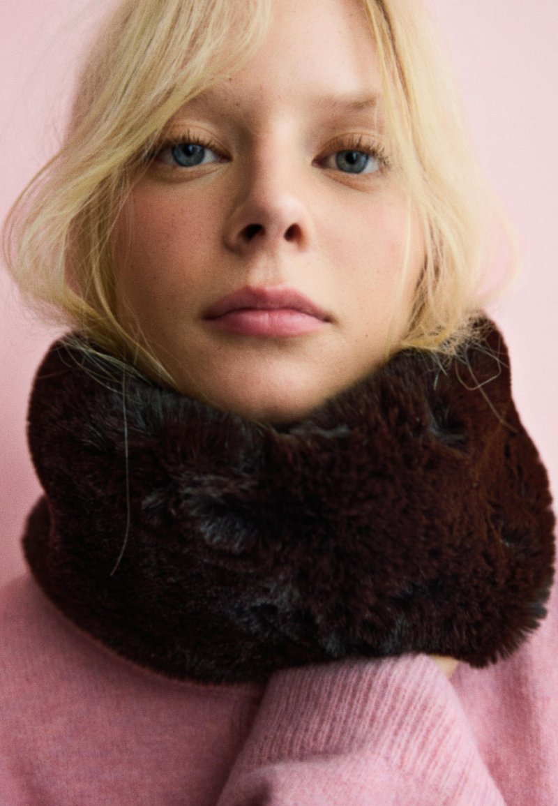 Col montant en fausse fourrure marron avec une texture douce, porté sur un pull rose clair. L'arrière-plan est d'une couleur rose uni.