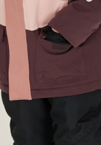 Vadderad jacka i rosa och burgundyröd med dragkedjsstängning, som har framfickor och kardborreaccenter. Tyget har en slät yta.
