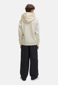 Un enfant aux cheveux bouclés porte un sweat à capuche beige clair et un pantalon large noir, se tenant de dos face à un fond uni.