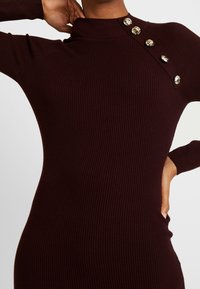 Robe côtelée bordeaux avec un col montant, featuring des détails de boutons en argent sur l'épaule et des manches longues ajustées. Tissu doux et texturé.