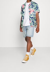 Chemise à manches courtes à motif floral avec des fleurs roses sur un fond bleu marine, associée à un short en denim bleu clair et des baskets montantes jaunes.