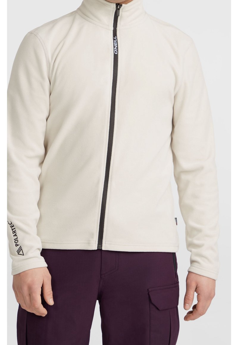 Homme portant une veste polaire crème zippée avec une fermeture éclair noire et un pantalon cargo violet, debout devant un fond uni.