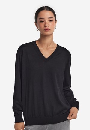 Kvinde med mørkt hår trukket tilbage iført en løs sort V-halset trøje med lange ærmer og lysegrå jeans, stående mod en ensfarvet baggrund.