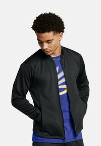 Black zip-front jacket with a smooth texture, raglan sleeves, and a fitted design; worn over a blue shirt featuring a graphic print.