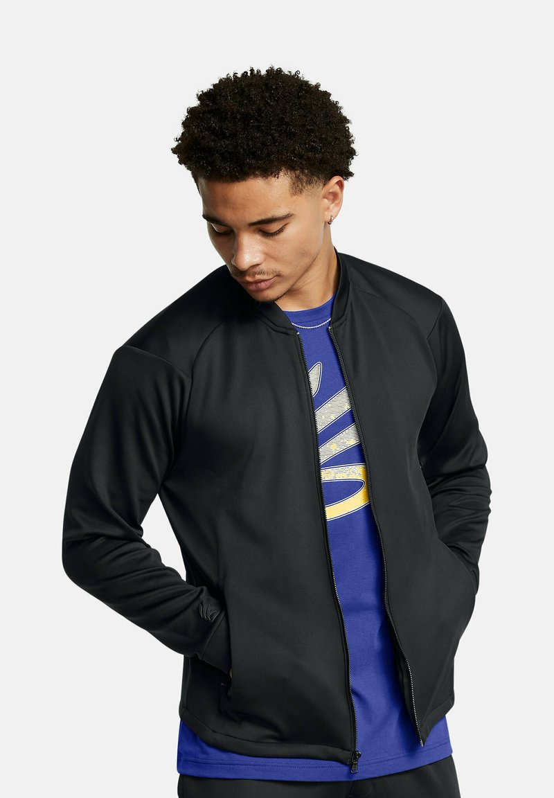 Black zip-front jacket with a smooth texture, raglan sleeves, and a fitted design; worn over a blue shirt featuring a graphic print.