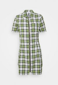 Vestido de manga corta a cuadros en verde, blanco y negro, con cremallera frontal, bolsillo en el pecho y detalle de cadena plateada sobre fondo blanco.