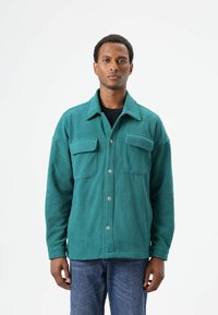 Turkooizen fleecejack met twee borstzakken en drukknopen, voorzien van een klassieke kraag en een losse pasvorm, gedragen met een blauwe spijkerbroek.