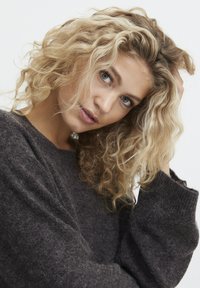 Mørkegrå struktureret sweater med brede ærmer, båret af en person med krøllet blond hår, der vender lidt til siden mod en hvid baggrund.