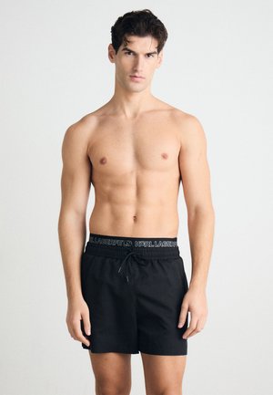 Shorts de bain noirs avec une taille élastique arborant un logo. Le tissu semble lisse, léger et a une coupe décontractée.
