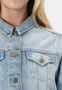 Light blue denim jacket with two chest pockets, gold-button accents, and a classic collar. Features faded texture and subtle stitching details.