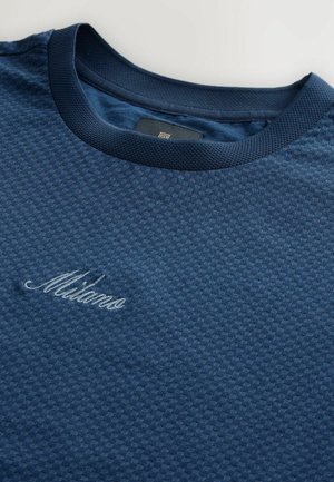 Tumed sinine tekstuuriga T-särk ümarate kaelusega ja ribaga kaeluses. Omab heledama sinise niidiga brodeeritud "Milano".
