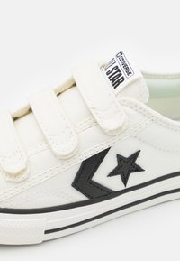 White canvas sneaker with three Velcro straps, black star logo, and contrasting black chevron design on the side. Rubber sole with stripes.
