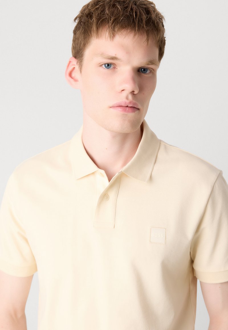 Polohemd aus leichtem gelben Baumwollstoff mit klassischem Kragen, kurzen Ärmeln und einem strukturierten Logo-Patch auf der Brust. Glatter Stoff.