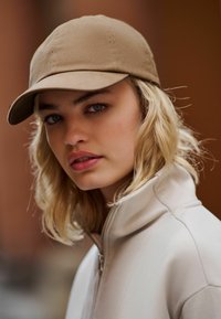 Cappellino tan realizzato in tessuto morbido con visiera curva, abbinato a una giacca leggera con zip, caratterizzata da un colletto alto e una texture liscia.