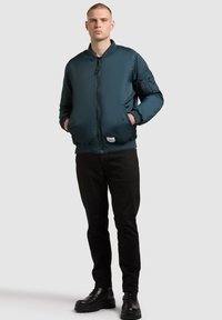 Ung man som står med händerna i fickorna, iklädd en tealgrön bomberjacka, svarta byxor och svarta snörskor mot en enkel bakgrund.