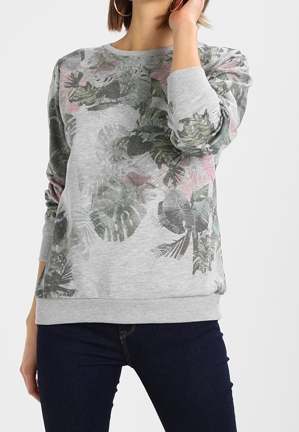 Femme portant un sweat-shirt gris avec des motifs de feuilles tropicales vertes et roses, et un jean bleu foncé, posant avec une main près du visage.