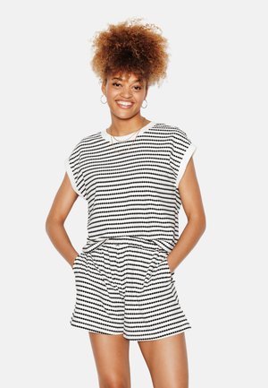 Jeune femme souriante, portant un top à manches courtes rayé noir et blanc et un short assorti, les mains dans les poches, cheveux bouclés relevés et boucles d'oreilles créoles.