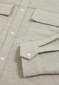 Cămașă ușor gri, texturată, cu guler, dotată cu două buzunare pe piept, butoane albe și detaliu de manșetă pliată. Materialul pare moale și călduț.