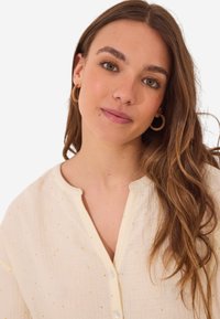 Crèmekleurige blouse met een knoopsluiting, voorzien van een textuurstof en gouden stipaccenten in het geheel. Lange mouwen met een ontspannen pasvorm.