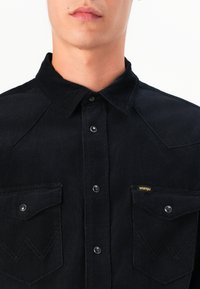 Camisa de pana negra con cuello puntiagudo, dos bolsillos en el pecho con botones a presión y un sutil acento de logo en uno de los bolsillos.