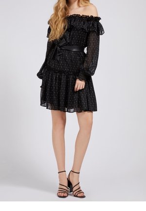 Frau in einem schwarzen, schulterfreien Kleid mit langen Ärmeln und weißen Punkten, tailliertem Gürtel und schwarzen, geschnürten High-Heel-Sandalen.