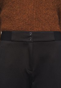 Pantaloni neri a trama con due bottoni rotondi neri in vita e tasche laterali, abbinati a un maglione lavorato a maglia marrone.