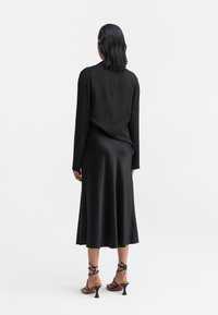 Sort satin lang kjole med lange ærmer, flydende nederkant og subtil glans. Sorte strop sandaler med små, tapered hæle komplementerer outfittet.