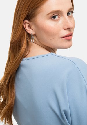 Helles blaues Oberteil aus glattem Stoff, mit einem runden Ausschnitt und Details an den Schulternähten. Kombiniert mit silbernen Ohrsteckern in Form von Ringen.