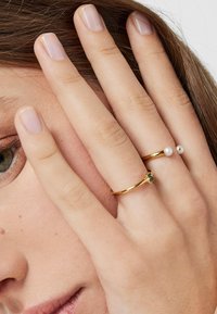 Dwa złote pierścionki na palcach: jeden ozdobiony małym zielonym akcentem, drugi białą perłą. Skóra ma naturalną teksturę; paznokcie są niepomalowane.