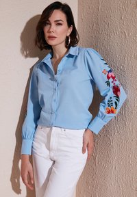 Light blue button-up shirt with puffed sleeves, featuring colorful floral embroidery on both sleeves. Paired with white jeans.