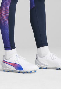 Scarpe da football bianche con dettagli blu e rosa, abbinate a leggings blu scuro con un motivo viola texturizzato.