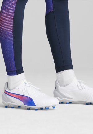 Scarpe da football bianche con dettagli blu e rosa, abbinate a leggings blu scuro con un motivo viola texturizzato.