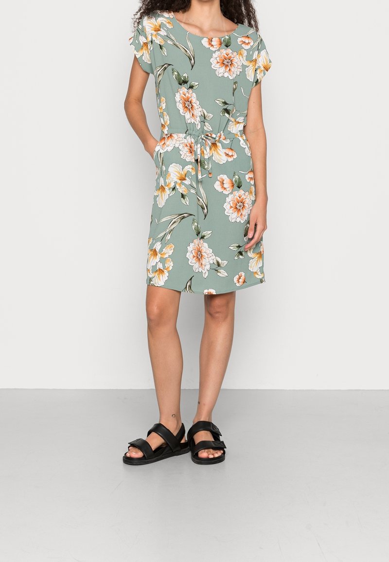 Robe florale en tissu vert avec des fleurs orange et blanches. Présente une coupe ample, des manches courtes et un lien à la taille ajustable. Sandales noires portées.