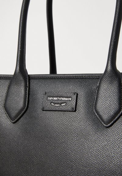 Geantă de mână din piele texturată neagră, cu două mânere, prezentând un plăcuță de marcă argintie pe care este inscripționat "Emporio Armani Milano 1981."